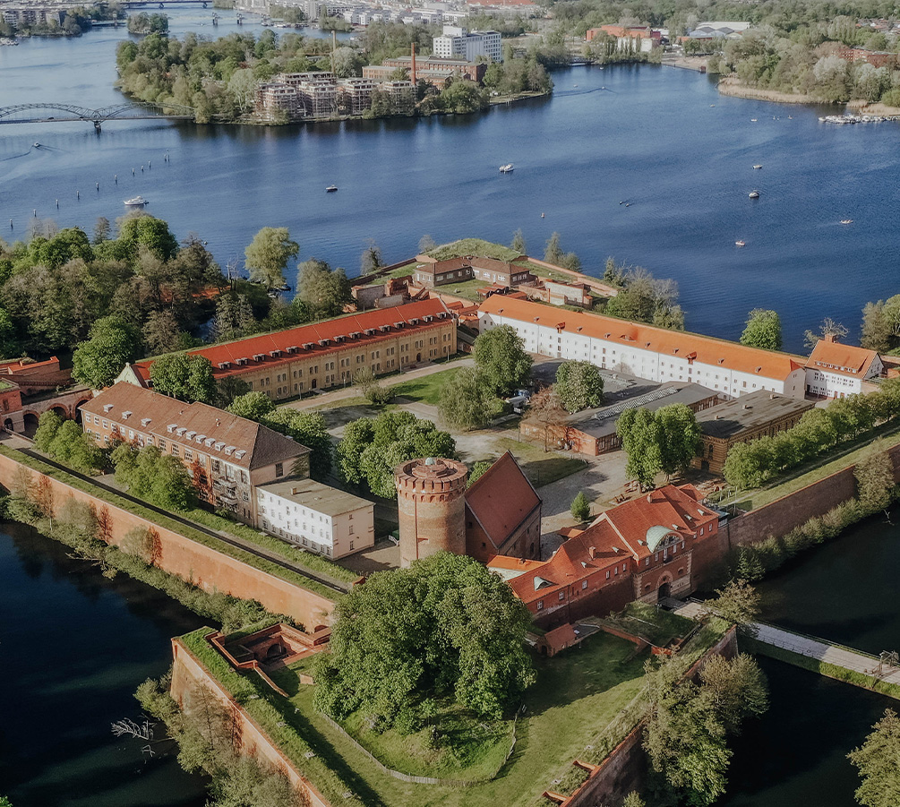 Luftaufnahme einer historischen Festungsanlage mit roten Ziegeldächern, umgeben von Wassergräben und Grünflächen, im Hintergrund ein Fluss mit Booten und Brücken.
